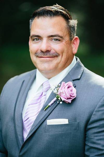 A handsome groom poses for his Groomsman wedding photo. He is wearing a grey fitted tuxedo with a pinstripe lavender neck tie that perfectly matches the lavender boutonniere with baby's breath and white ribbon. In folding his pocket handkerchief he chose to fold it into a white square that just peeks above the pocket on his grey wedding attire. 