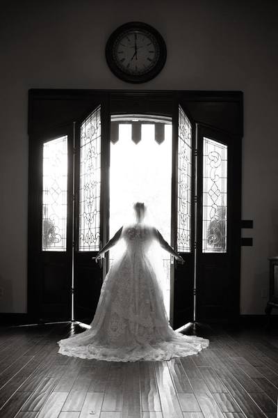 This is a timeless wedding photo. The Bride stands in open doors as she prepares to walk outside on the best day ever- her wedding. The leaded glass panes of these huge double doors show through to the Sterling Castle Knights standing sentry on either side of the wooden faux drawbridge. You can see the intricate details in her embroidered wedding veil and all of the patterns in her  bridal train as it falls perfectly around her. Her ballgown style wedding dress is very ornately detailed with floral stitching and a sweep train to give her dress a flowy and yet easy to move about in feel. 