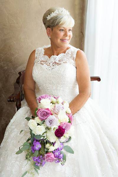 A Beautiful lady with a pixie bob wedding hairstyle stands in a beautifully decorated bridal suite. The lavish Bridal Suite has a hand brushed stucco bronze wall and soft sunshine floods through the sheer white drapery. The bride is wearing a jeweled tiara with pearls in her hair and she is holding a cascade style bouquet full of lavendar, pink, and white roses, purple stock orchids, seeded eucalyptus, white spray roses and lilies. Her stylish A-line wedding dress is modest with lace making up the shoulder straps and flowing down over most of the dress. 